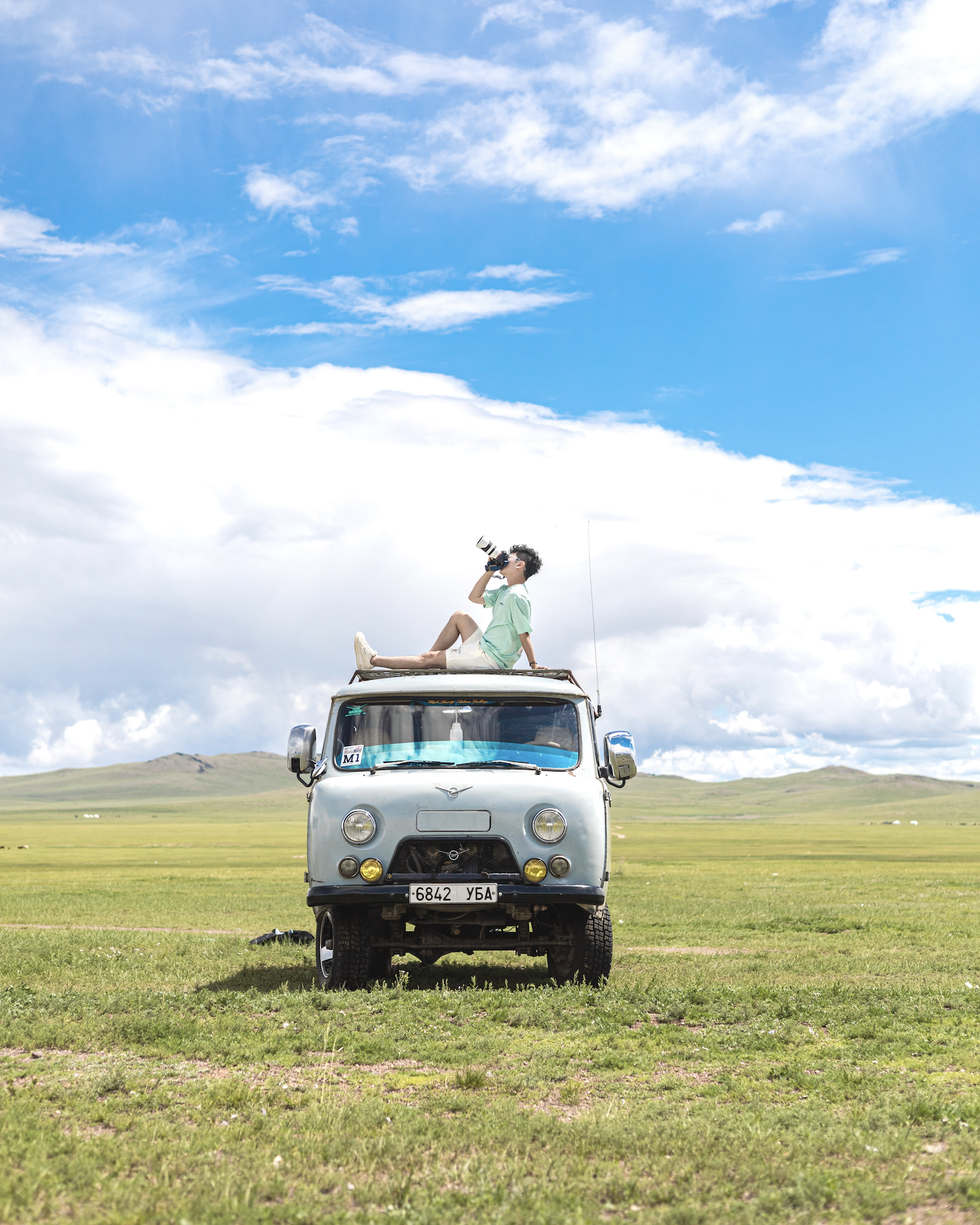 Man sitting on the roof of a vehicle in a field and holding a camera up to the sky