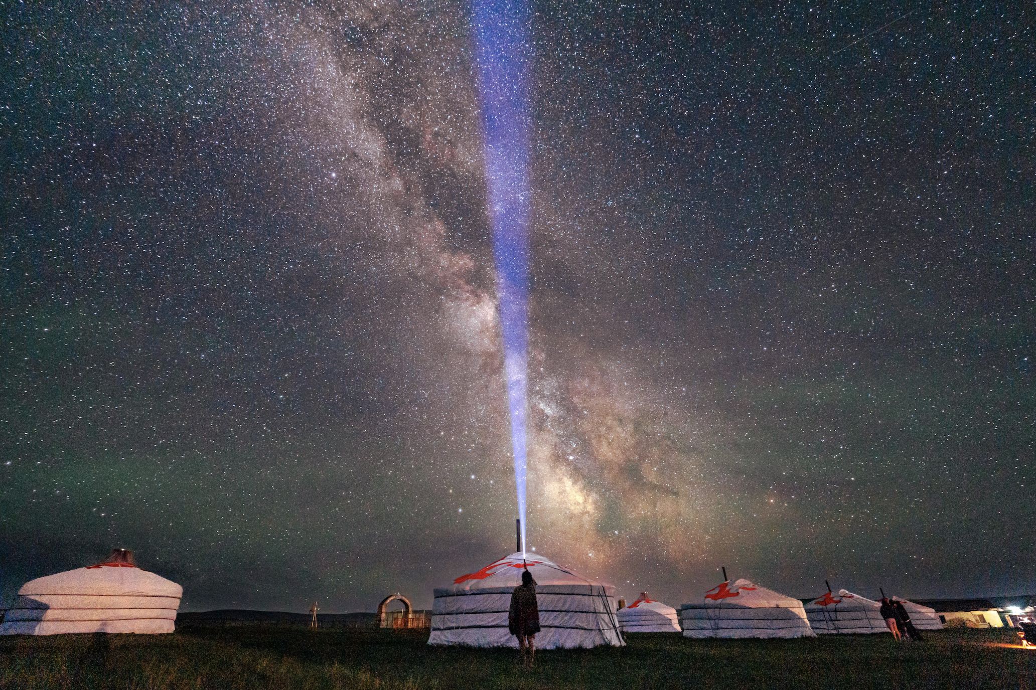 Person standing in front of a group of yurts and holding a flashlight pointing up at the starry, night sky 