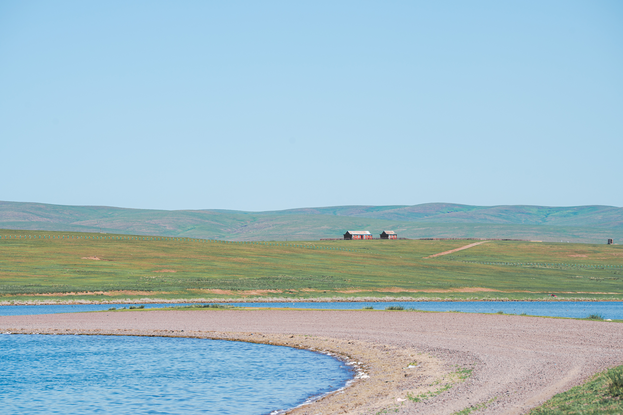 Wide shot of a vast landscape with a lake in the foreground and a house and fences at a distance