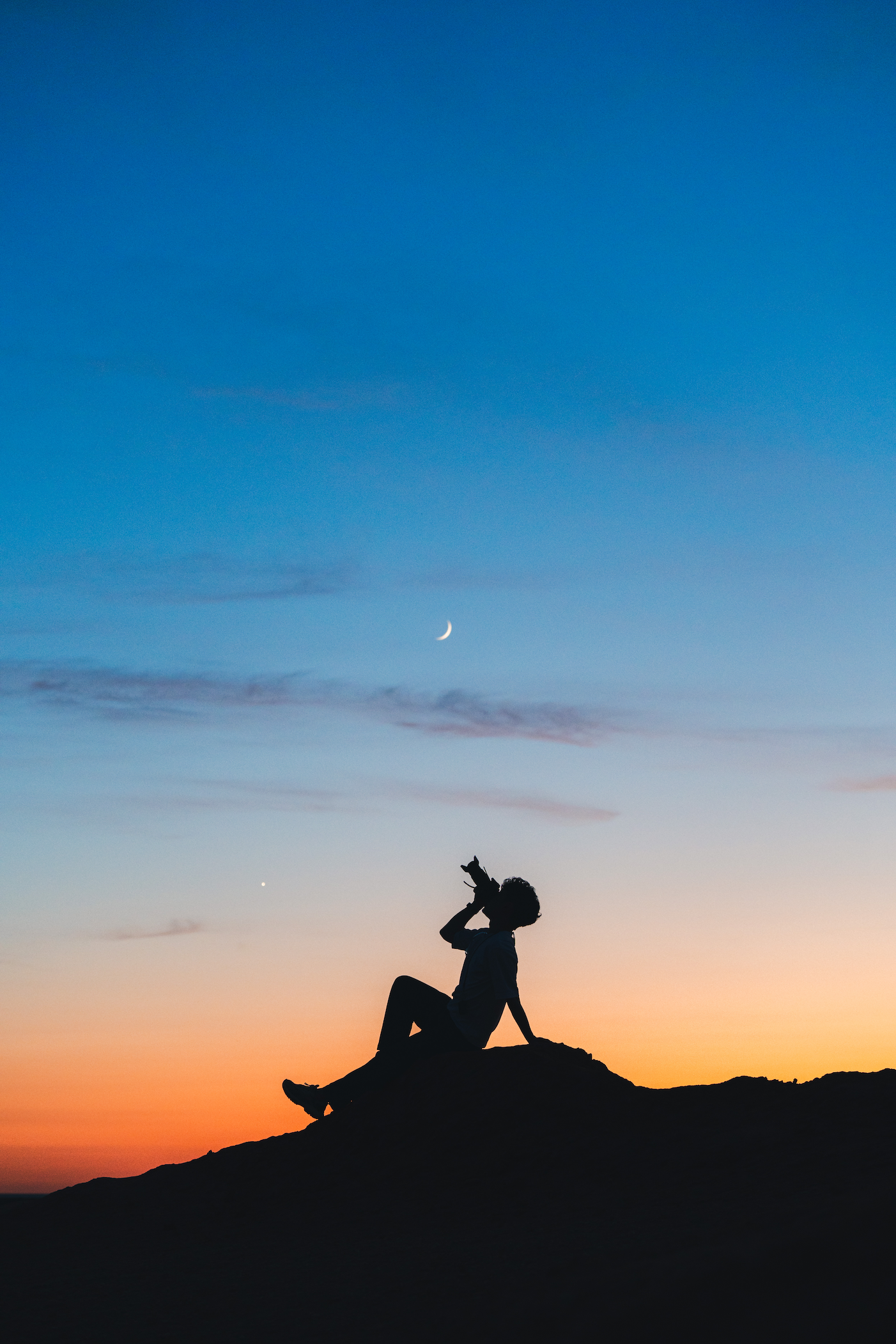 Silhouette of a man sitting on a rock at dusk and holding a camera up to the sky