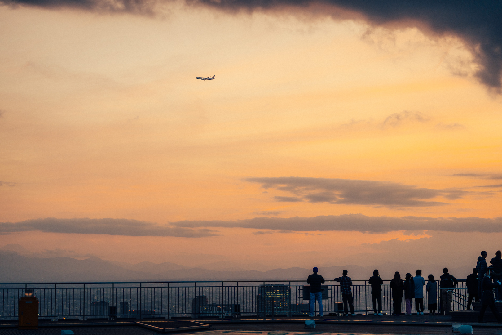 People watching the sunset on an observation deck with an airplane moving across the orange sky