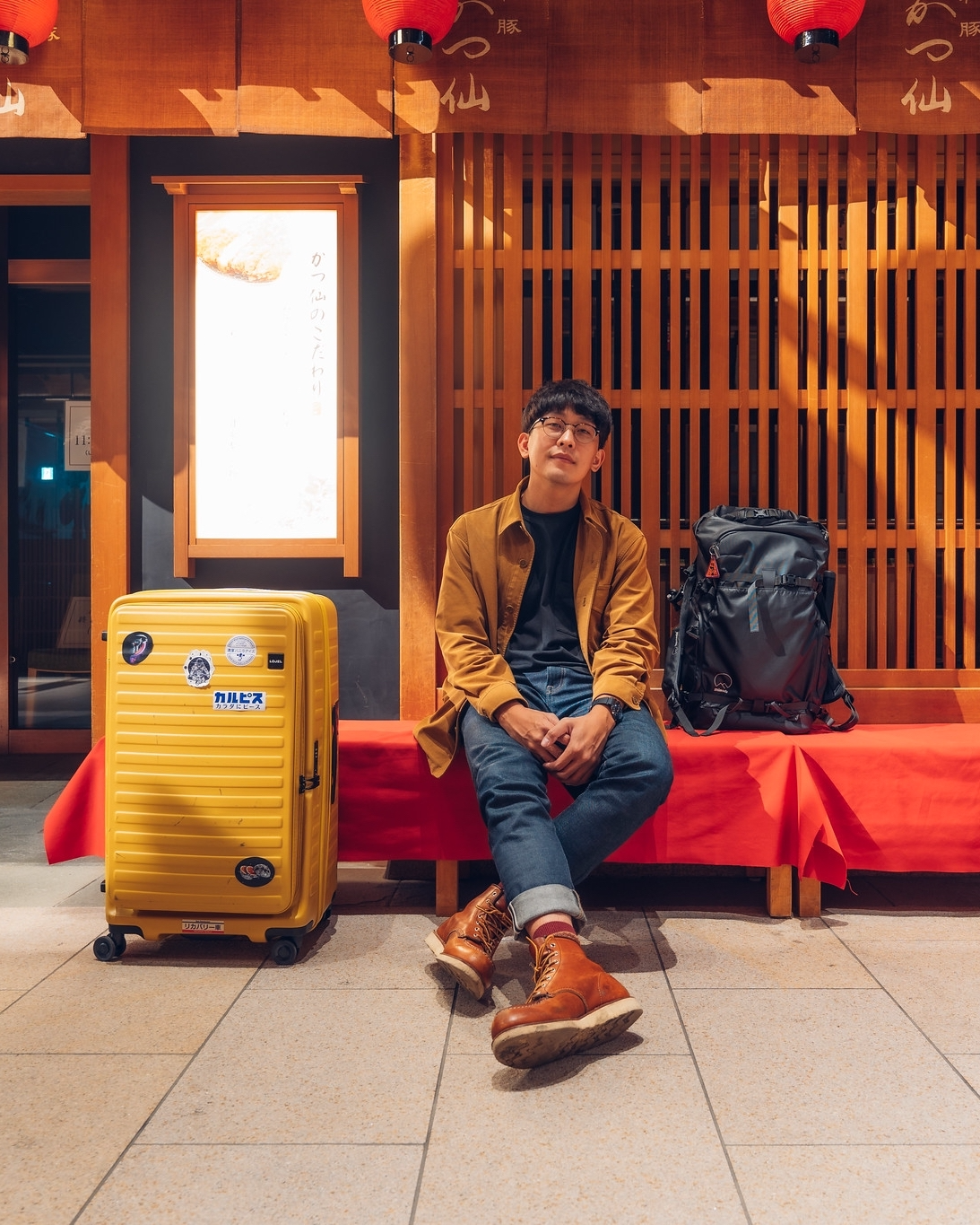 Man sitting on benches outside a store with his suitcase and backpack